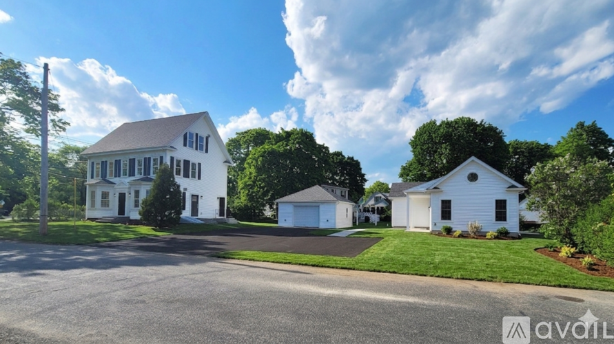 A white house with a brown roof is surrounded by greenery and a clear sky.