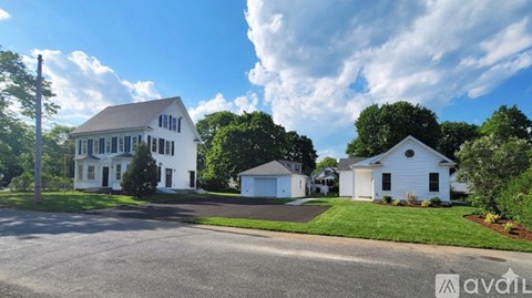 A white house with a brown roof is surrounded by greenery and a clear sky.