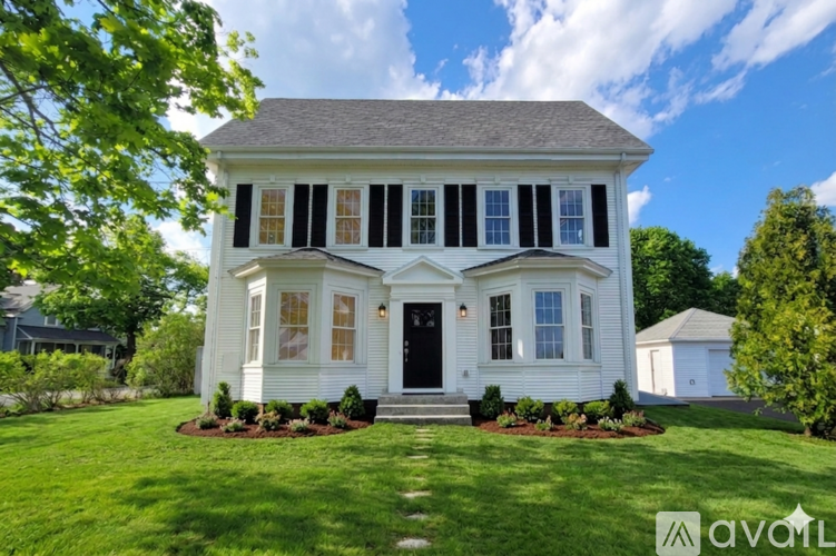 A white house with a black door and windows is surrounded by greenery.