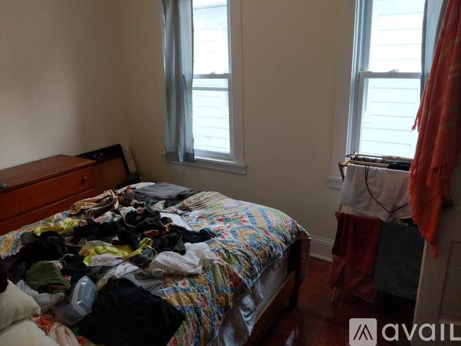 A messy bedroom with a bed covered in clothes and a window with a red curtain.