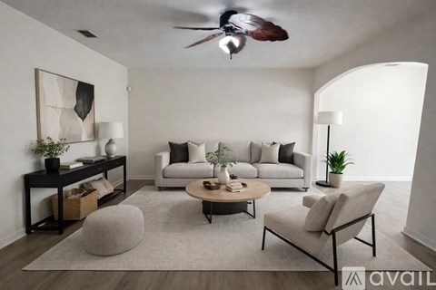 A living room with a grey sofa, a grey armchair, a wooden coffee table, a grey pouf, a wooden side table with a plant, a painting on the wall, and a ceiling fan.