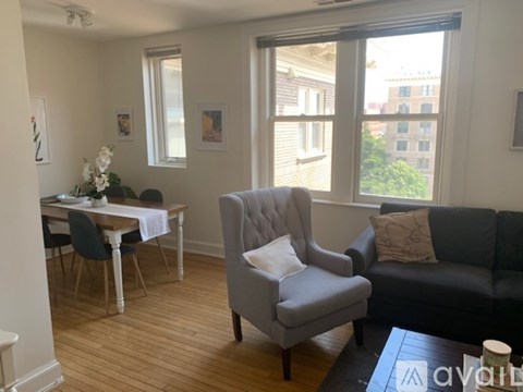 A living room with a grey armchair, a black sofa, a dining table with chairs, and a window with a view of the city.