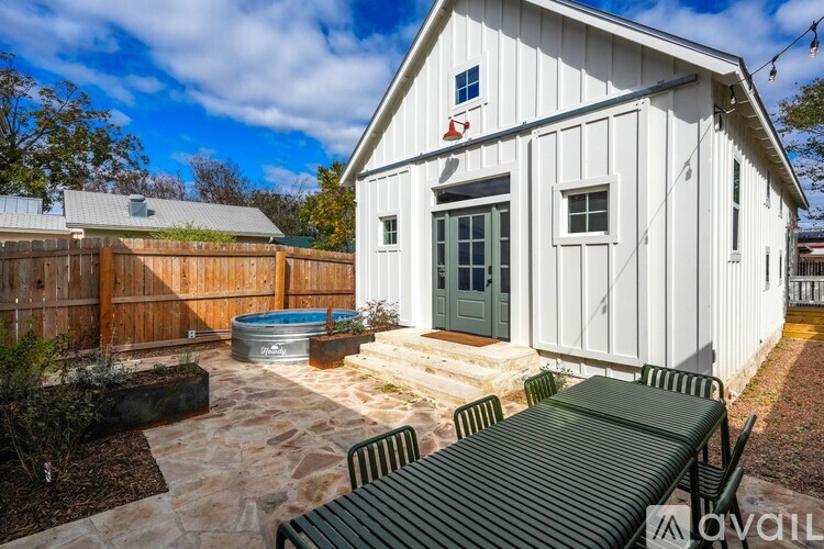 A white barn-like building with a green door and windows, a wooden fence, and a stone patio with a bench.