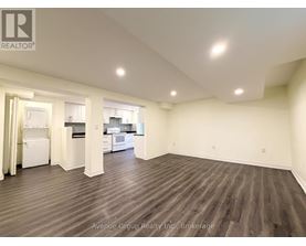 A large, empty room with wood flooring and a kitchen visible through the open door.