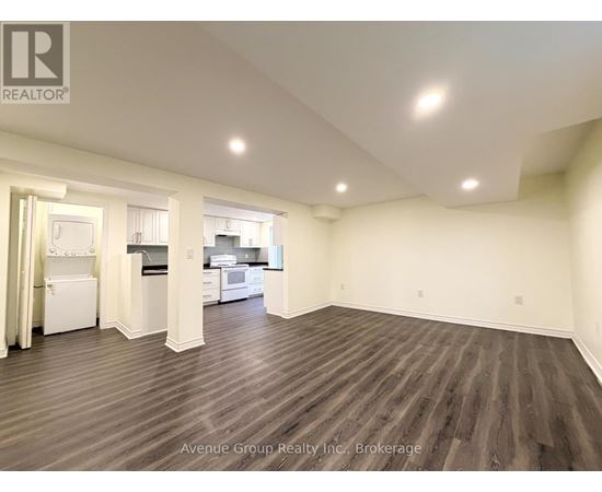 A large, empty room with wood flooring and a kitchen visible through the open door.