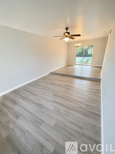 A room with a ceiling fan and light, wooden flooring, and a window with a view of greenery outside.