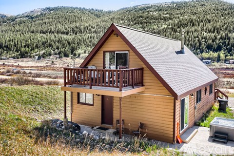 A small wooden cabin with a balcony is situated in a grassy area with trees in the background.