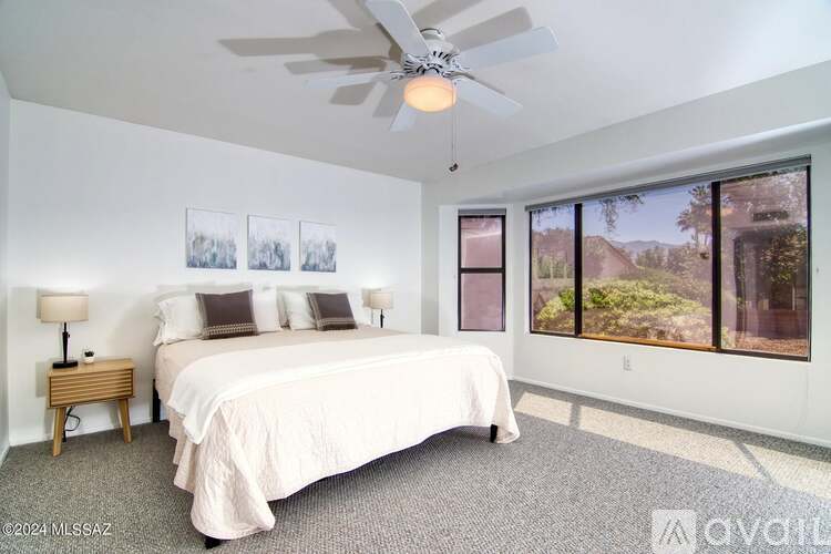 A bedroom with a large bed, a ceiling fan, and a view of the outdoors through the windows.