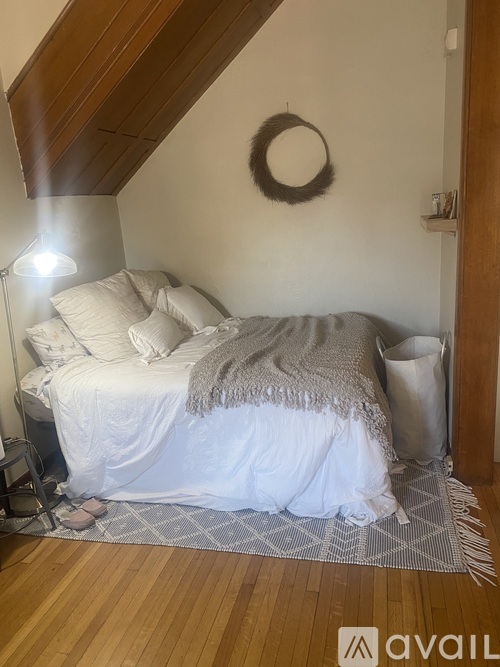 A bedroom with a bed covered in a white sheet and a grey blanket, a wreath on the wall, and a wooden floor.
