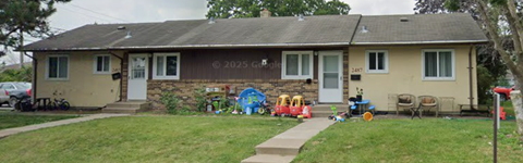 A house with a white door and windows is surrounded by a lawn and a sidewalk.