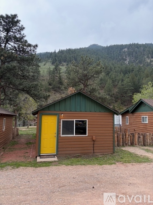 A small brown cabin with a yellow door is situated in a grassy area with a mountain in the background.