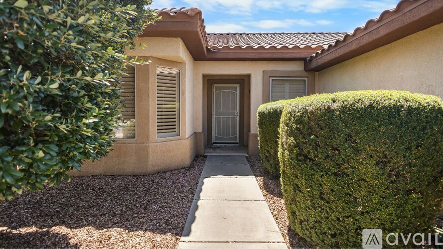 A house with a brown roof and tan walls has a small front yard with a hedge and a concrete path leading to the door.