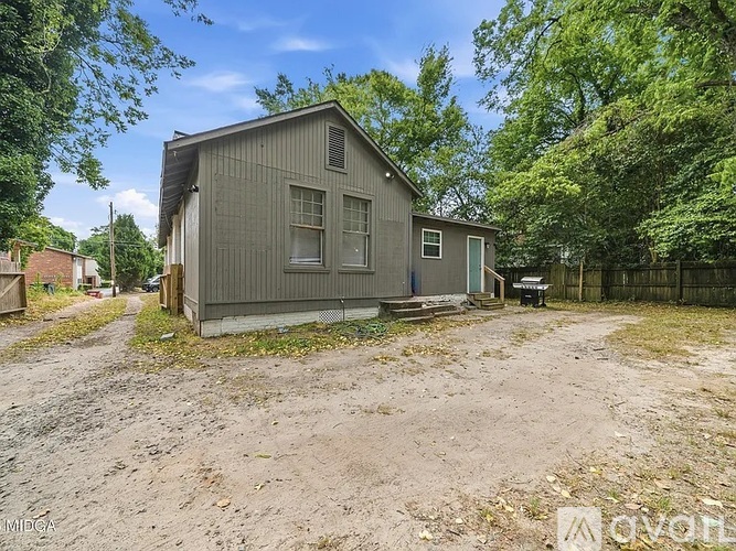 A grey house with a blue door is surrounded by trees and a dirt area.