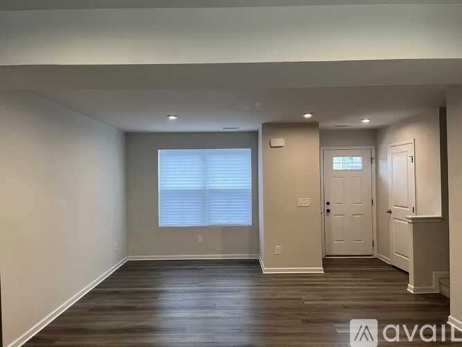A spacious room with wooden floors and white walls, featuring a window with blinds and a closed door.