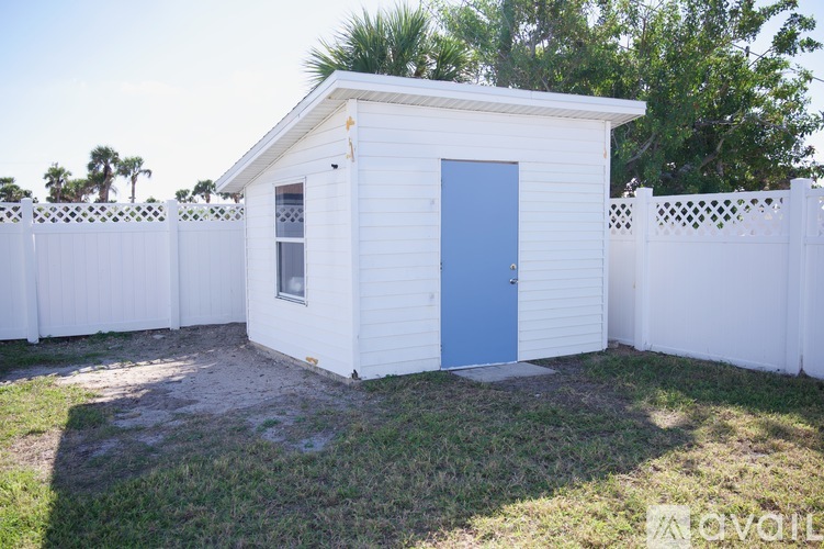 A small white shed with a blue door is surrounded by a white fence.