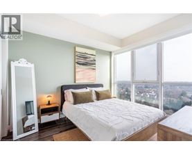 A bedroom with a large bed, a dresser, and a large window overlooking a cityscape.