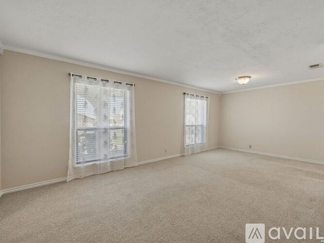 A room with beige walls and carpet, two windows with white curtains, and a ceiling with a light fixture.