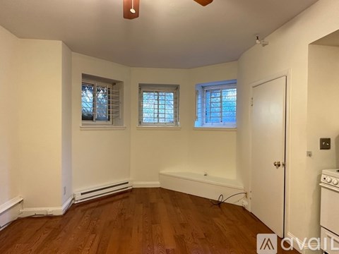 A room with wooden floors and white walls, featuring a window with blinds, a door, and a small stove.