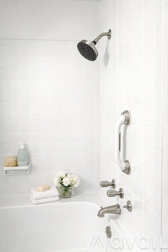 A white tiled bathroom with a shower head, faucet, and a small shelf with toiletries.