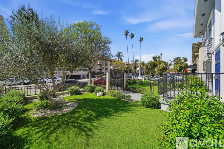 A well-maintained garden with a tree, shrubs, and a lawn in the foreground, with a parking lot and buildings in the background.
