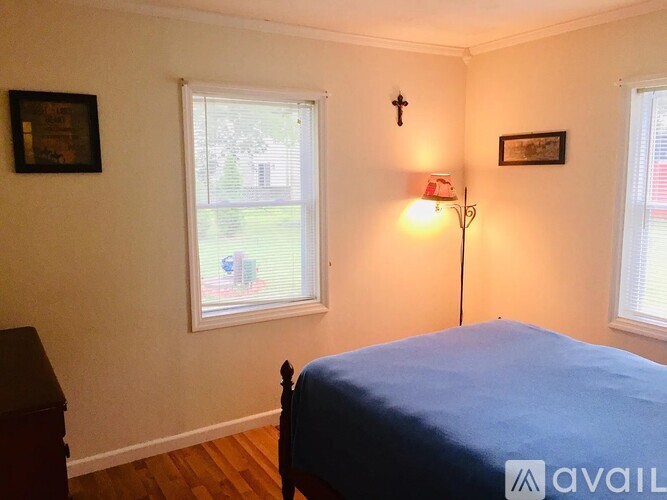 A bedroom with a bed, a cross on the wall, and a window with a view of a lawn and a house.