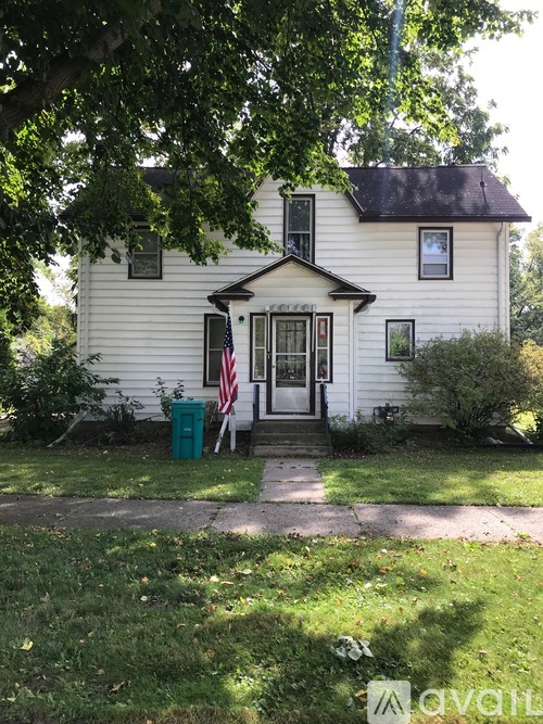 A white house with a green trash can and an American flag on the front porch.