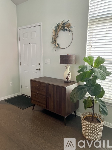 A room with a white door, a wreath on the wall, a wooden cabinet, a lamp, and a plant.