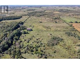 An aerial view of a large, open field with a small house in the distance and a forested area to the left.