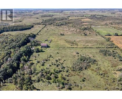 An aerial view of a large, open field with a small house in the distance and a forested area to the left.