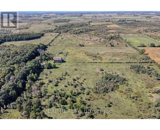 An aerial view of a large, open field with a small house in the distance and a forested area to the left.