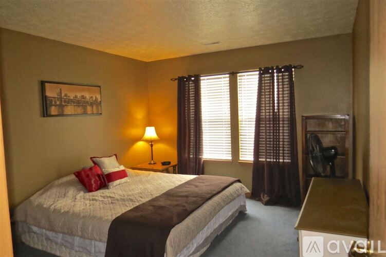 A bedroom with a bed, a nightstand, a lamp, a fan, and a window with curtains.