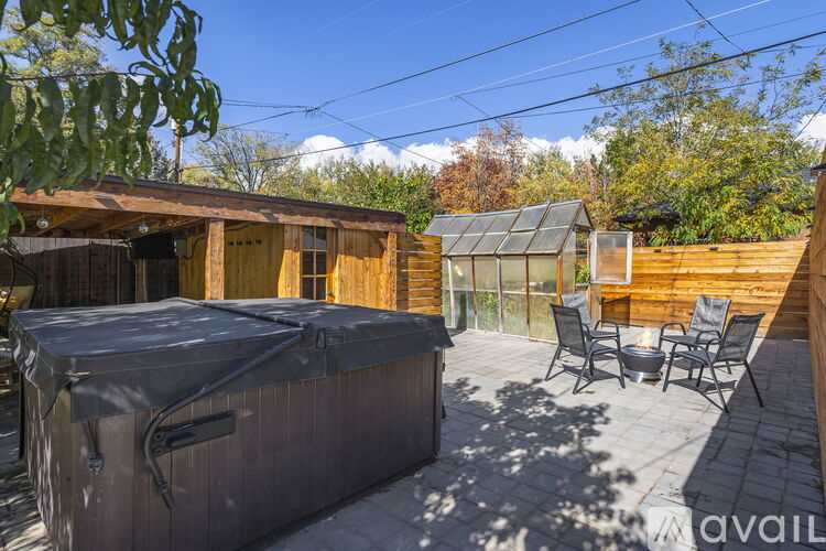 A hot tub sits on a patio with a wooden deck and a green tree in the background.