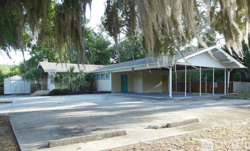 A building with a white roof and a covered porch is surrounded by trees with Spanish moss hanging from them.