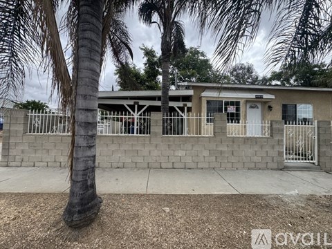 A house with a white door and windows is surrounded by a fence and a palm tree.