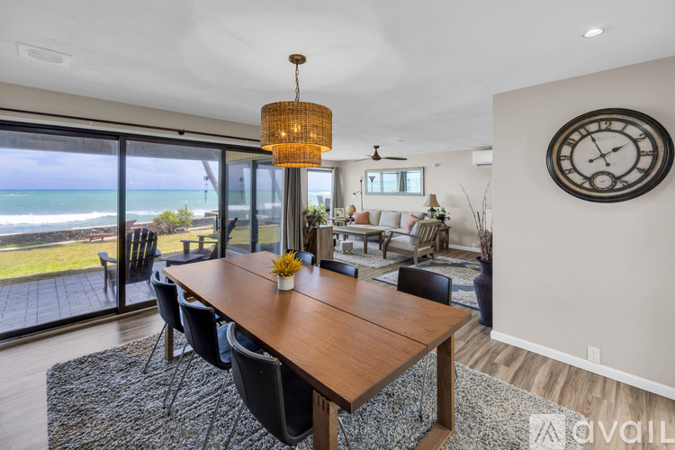 A dining room with a wooden table and chairs, a ceiling fan, and a large window overlooking the ocean.