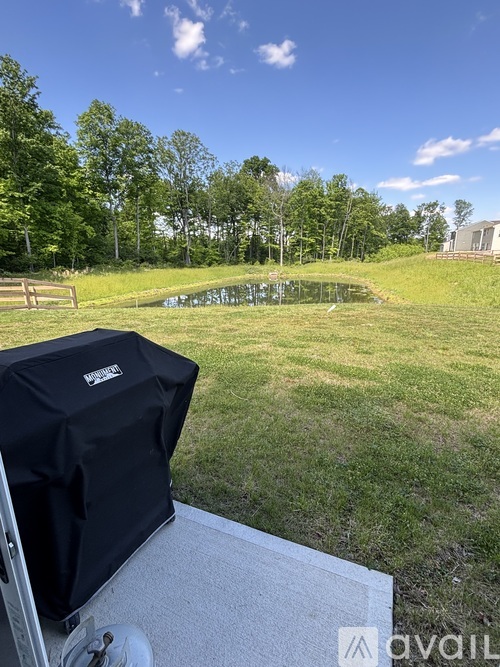 A BBQ grill is on a patio overlooking a grassy field and a pond.