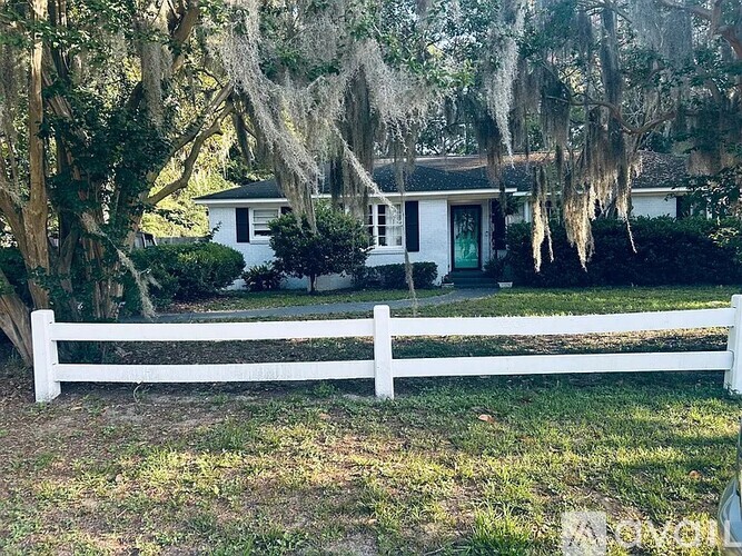 A white house with a white fence and trees with Spanish moss hanging from them.