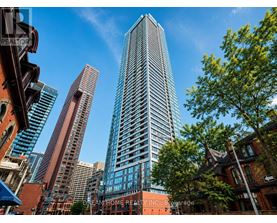 A tall building with many windows stands in front of a tree and other buildings.