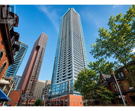 A tall building with many windows stands in front of a tree and other buildings.