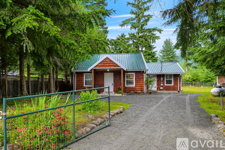 A house with a green roof is surrounded by trees and has a gravel driveway leading to the front door.