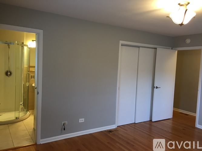A room with a wooden floor and white walls, with a door on the right and a glass shower area on the left.