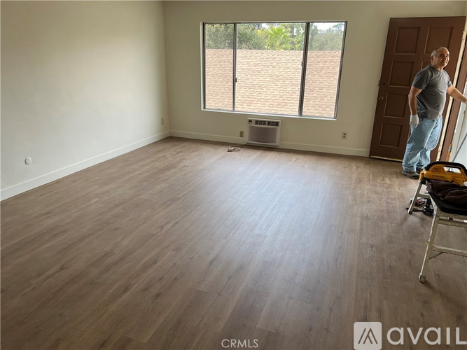 A man is standing in a room with wooden flooring and a window with blinds.