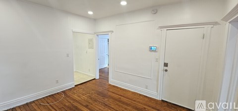 A room with white walls and wooden floors, with a door on the right and a window in the distance.