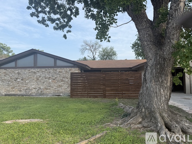 A house with a brown roof and a stone wall is surrounded by green grass and trees.