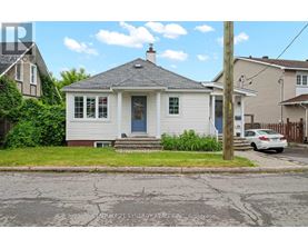 A small white house with a grey roof and a blue door is situated on a street with a cracked asphalt surface.