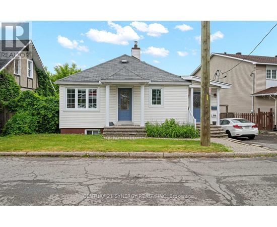 A small white house with a grey roof and a blue door is situated on a street with a cracked asphalt surface.