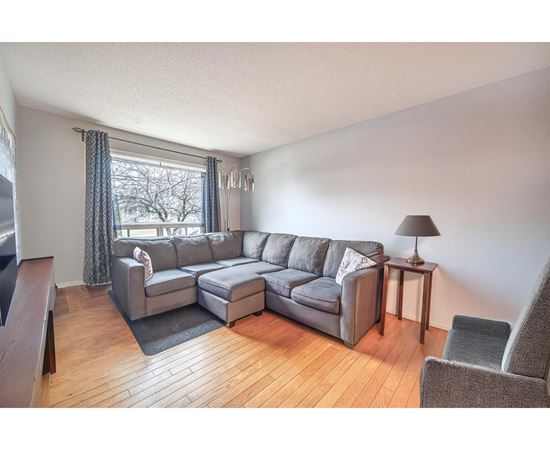A grey sectional sofa with a chaise lounge and ottoman in a living room with a wooden floor and a window with curtains.