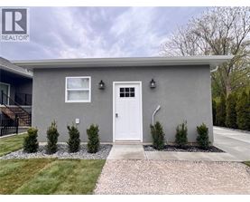A grey house with a white door and windows, surrounded by a gravel path and greenery.