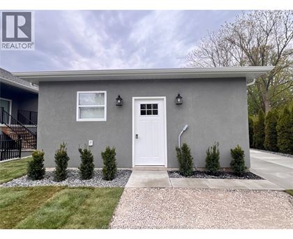 A grey house with a white door and windows, surrounded by a gravel path and greenery.