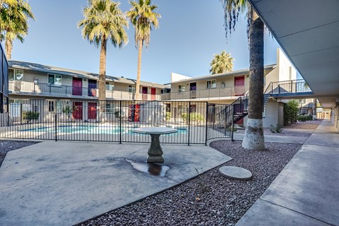 A concrete patio with a table and chairs is surrounded by a black fence and palm trees.